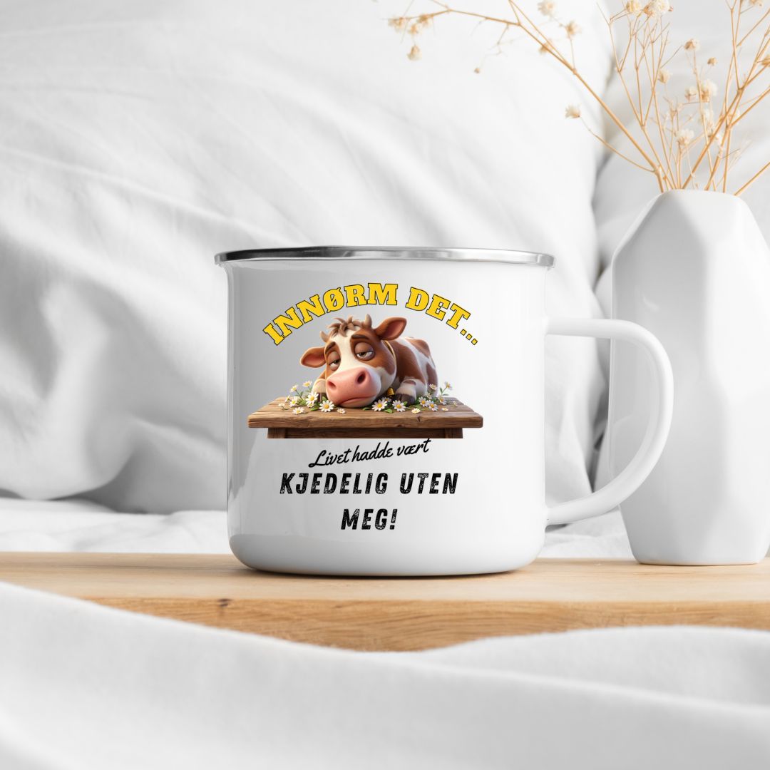 Hvitt emaljekrus med sølvkant og motiv av en brun og hvit ku som hviler hodet på et trebord med blomster, samt teksten Innrøm det... Livet hadde vært kjedelig uten meg!, plasseres på et trebord foran en hvit pute