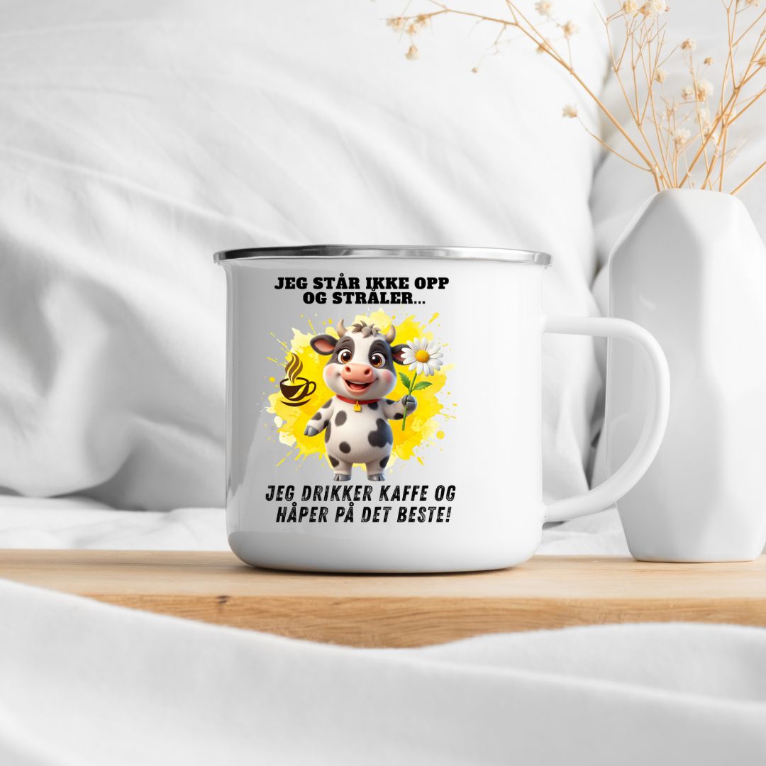 Hvitt emaljekrus med sølvkant og motiv av en hvit og sort ku som holder en margeritt foran gul fargesprut og en kaffekopp, med teksten Jeg står ikke opp og stråler... Jeg drikker kaffe og håper på det beste!, stående på et trebord